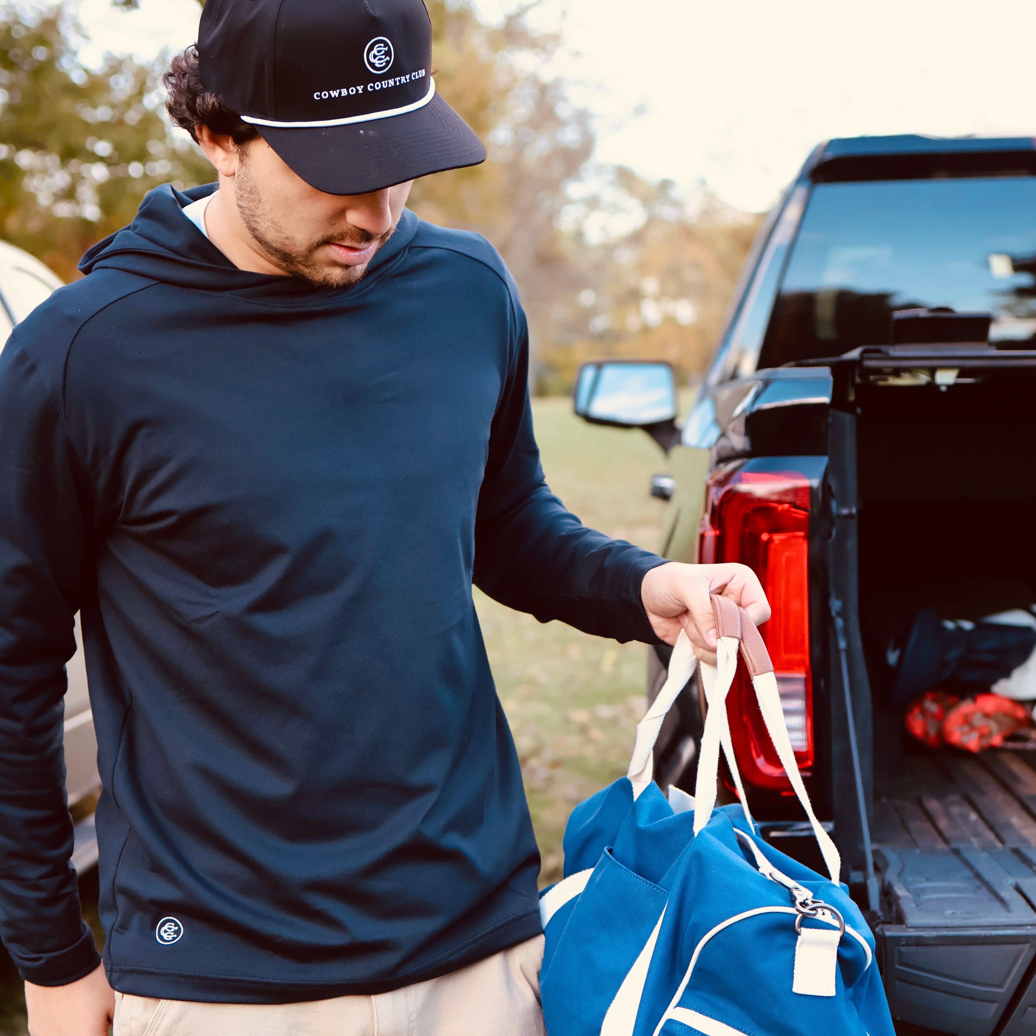 Man in navy hoodie and cap holding a blue duffel bag near an open truck bed outdoors.